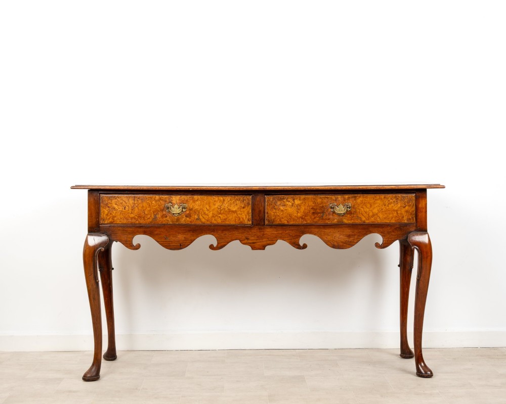 A Queen Anne style burr walnut console table featuring two drawers, a shaped apron, and four slender cabriole legs terminating in pad feet.