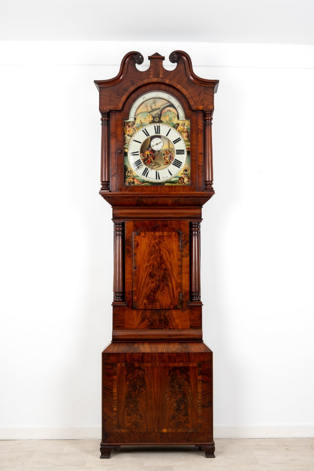 An antique mahogany longcase clock featuring a swan-neck pediment, turned columns, and a hand-painted arch dial with a moon phase display.