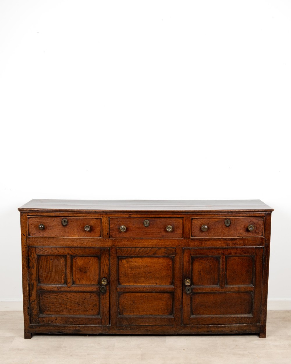 A Georgian antique oak dresser base featuring three upper drawers with metal keyhole escutcheons above three panelled cupboard doors. View IV