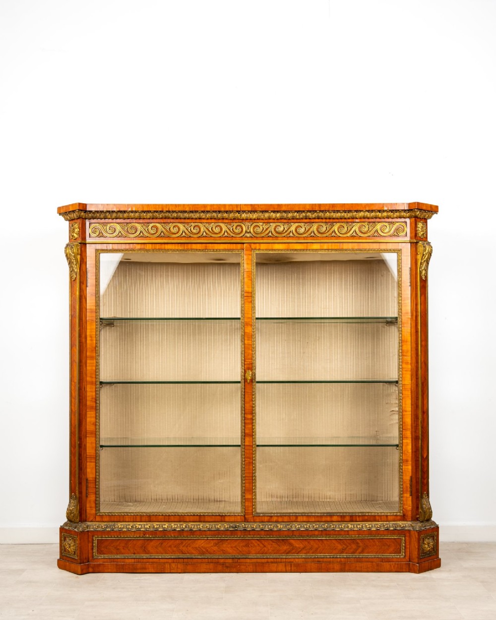 A large veneered display cabinet featuring gilt-bronze mounts, glazed twin doors, glass shelving, and a fabric-lined interior.