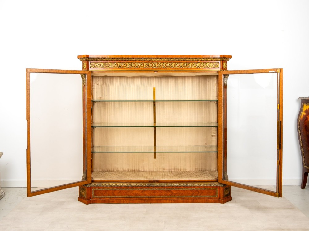 A large veneered display cabinet featuring gilt-bronze mounts, glazed twin doors, glass shelving, and a fabric-lined interior. View X