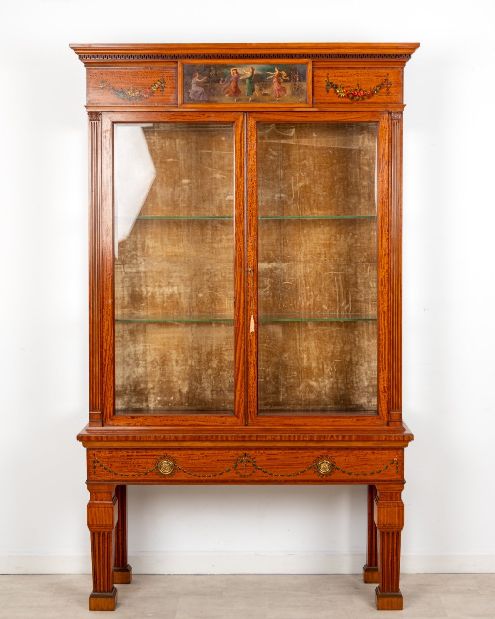 An antique Edwardian painted satinwood display cabinet featuring two glazed doors, glass shelves, fluted tapering legs, and classical hand-painted figurative and floral panels.