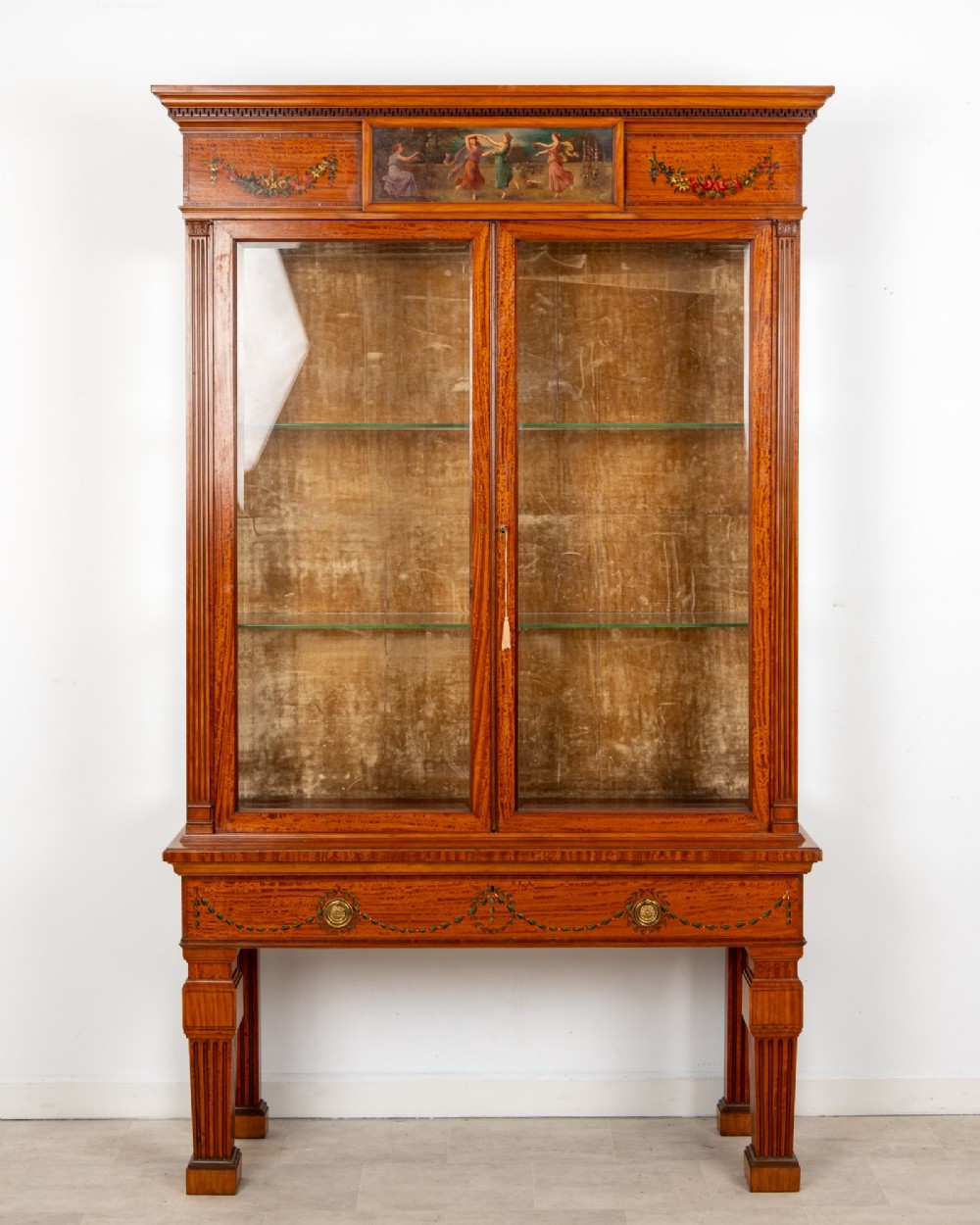 An antique Edwardian painted satinwood display cabinet featuring two glazed doors, glass shelves, fluted tapering legs, and classical hand-painted figurative and floral panels. View II