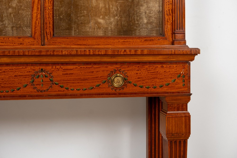 An antique Edwardian painted satinwood display cabinet featuring two glazed doors, glass shelves, fluted tapering legs, and classical hand-painted figurative and floral panels. View IV