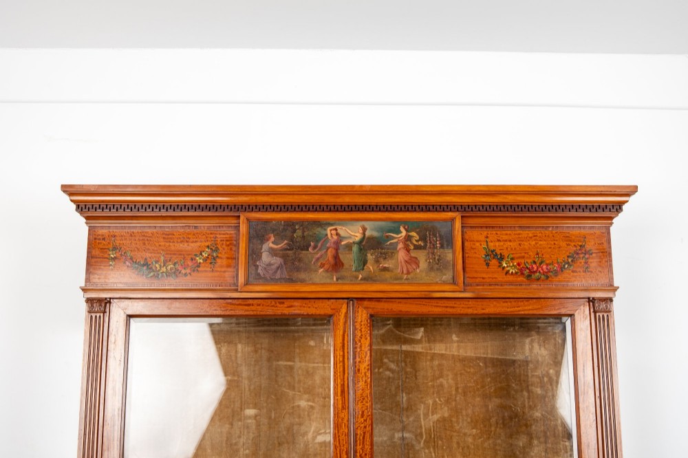 An antique Edwardian painted satinwood display cabinet featuring two glazed doors, glass shelves, fluted tapering legs, and classical hand-painted figurative and floral panels. View VI