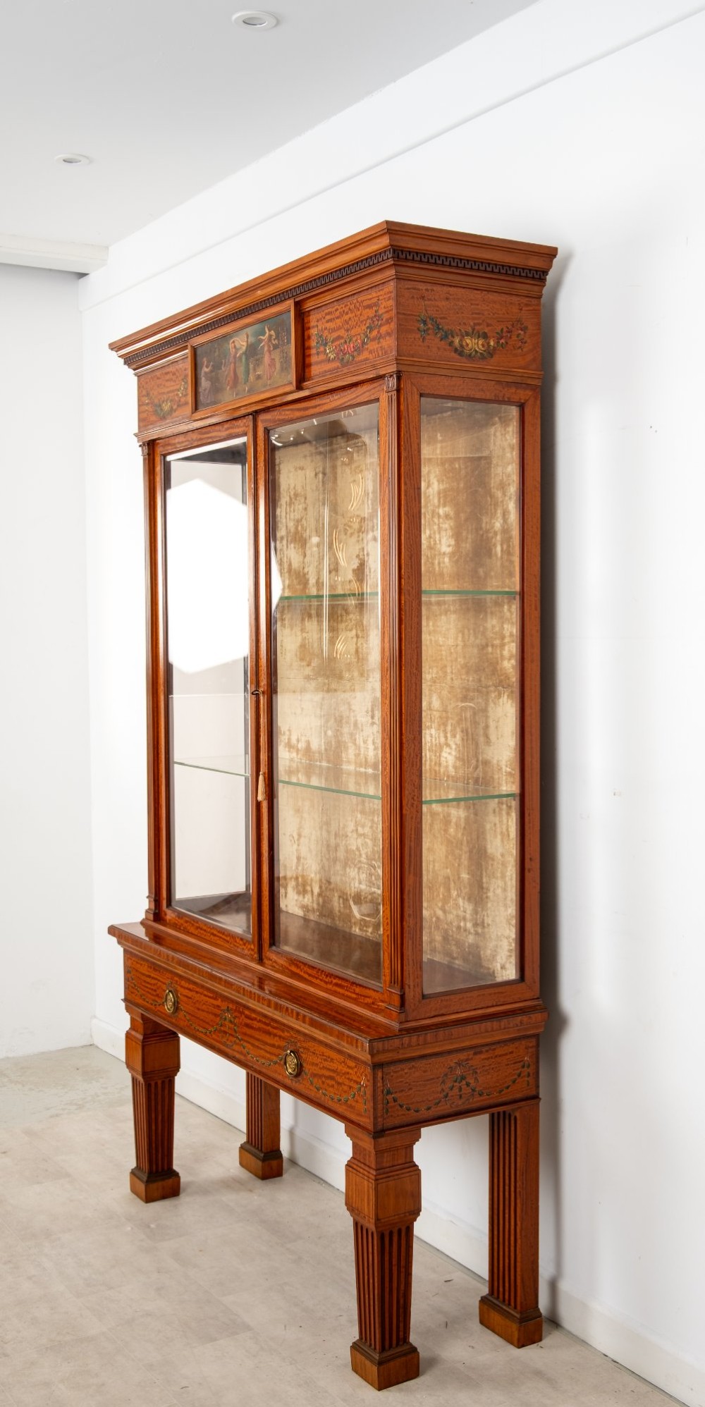 An antique Edwardian painted satinwood display cabinet featuring two glazed doors, glass shelves, fluted tapering legs, and classical hand-painted figurative and floral panels. View VII