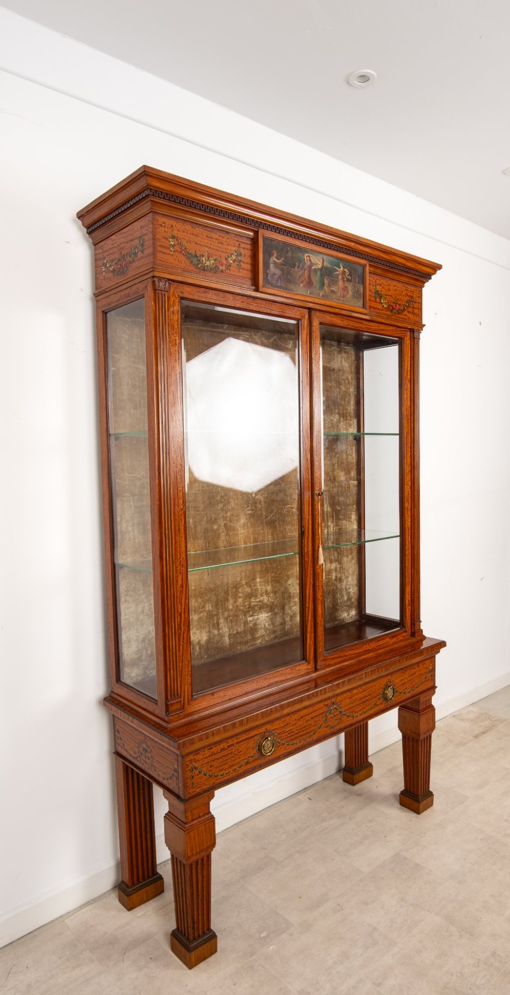 An antique Edwardian painted satinwood display cabinet featuring two glazed doors, glass shelves, fluted tapering legs, and classical hand-painted figurative and floral panels. View VIII
