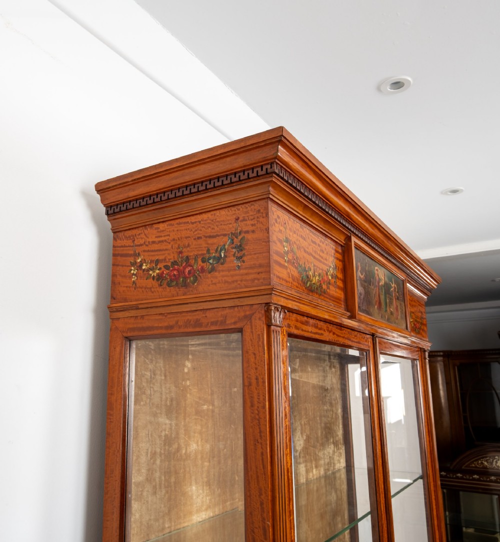 An antique Edwardian painted satinwood display cabinet featuring two glazed doors, glass shelves, fluted tapering legs, and classical hand-painted figurative and floral panels. View IX