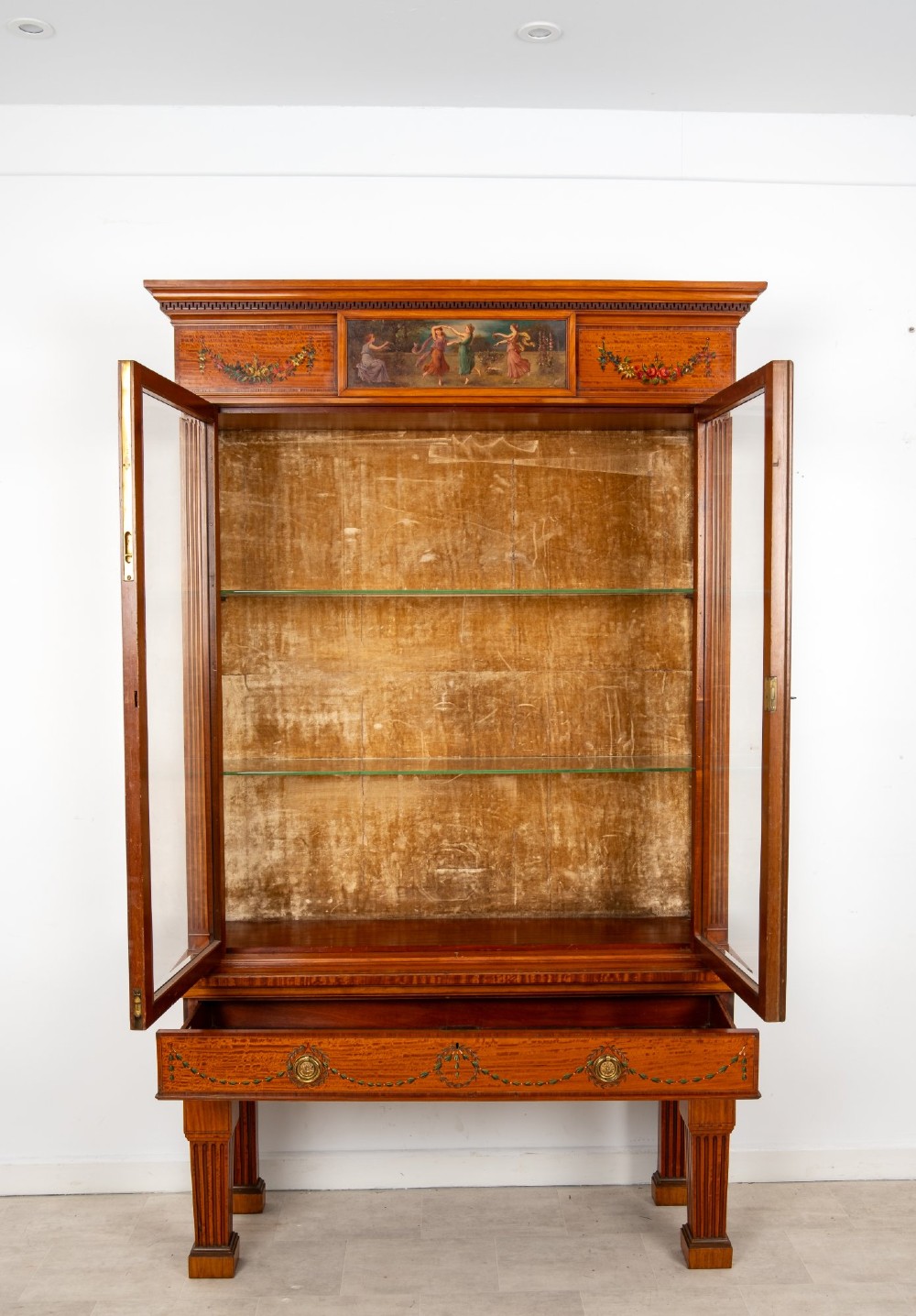An antique Edwardian painted satinwood display cabinet featuring two glazed doors, glass shelves, fluted tapering legs, and classical hand-painted figurative and floral panels. View XI