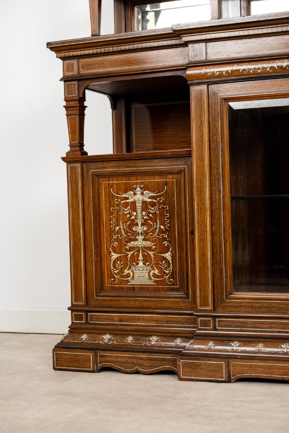 A large late Victorian rosewood display cabinet featuring intricate inlay work, glazed upper and lower doors, an arched pediment, and mirrored back panels. View VI