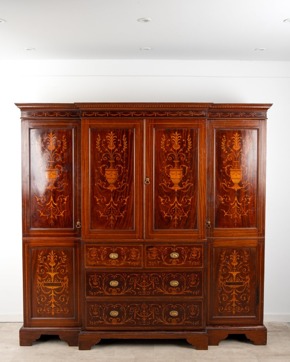 An Edwardian mahogany compactum wardrobe featuring classical urn marquetry inlay, side hanging sections, central drawers, and panelled cupboard doors. View II
