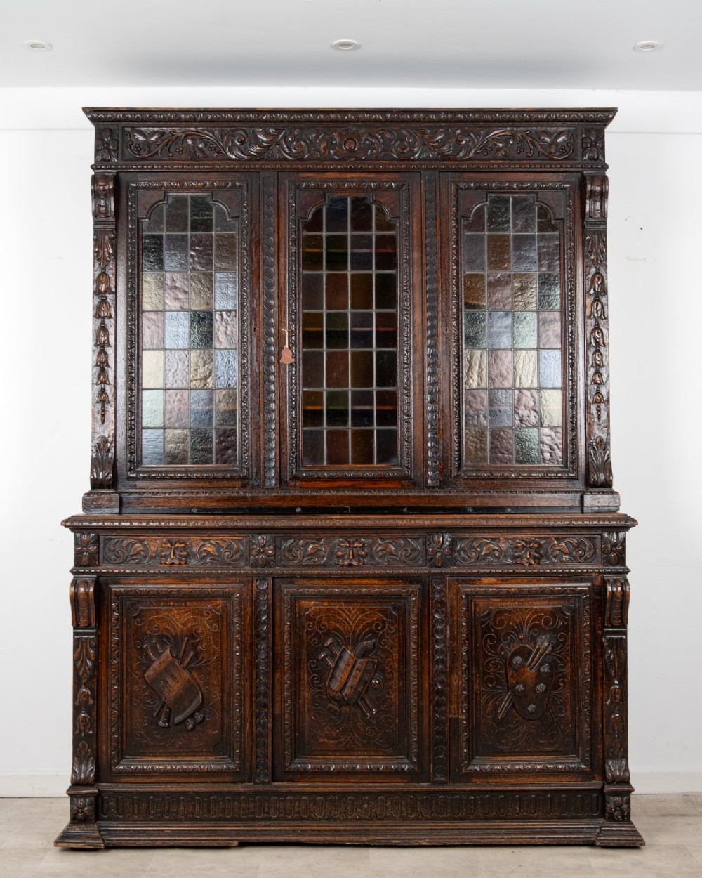 A large late Victorian dark carved oak bookcase featuring three upper glazed doors with coloured leaded glass panels above a lower cupboard base with relief carved emblems.