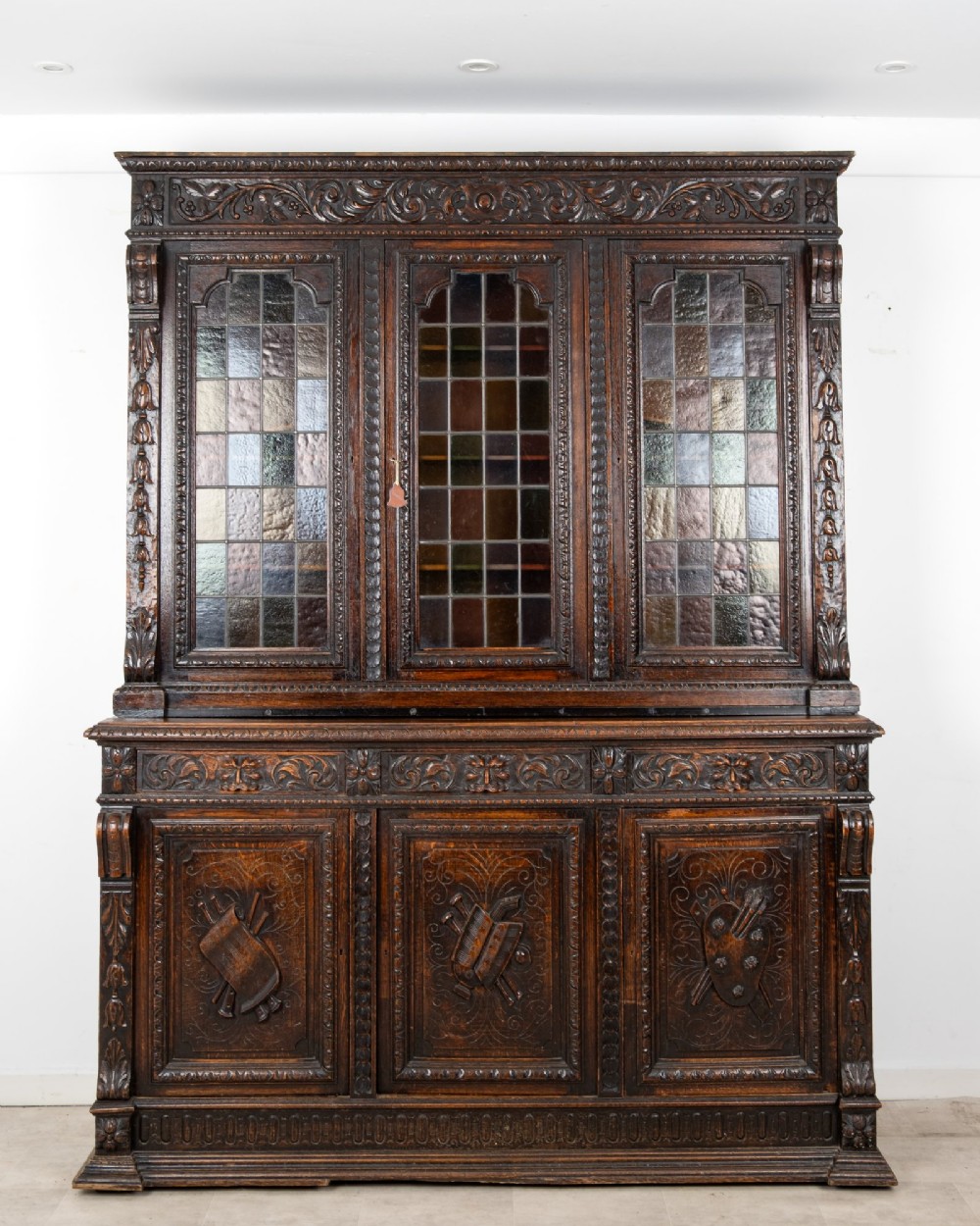 A large late Victorian dark carved oak bookcase featuring three upper glazed doors with coloured leaded glass panels above a lower cupboard base with relief carved emblems. View II