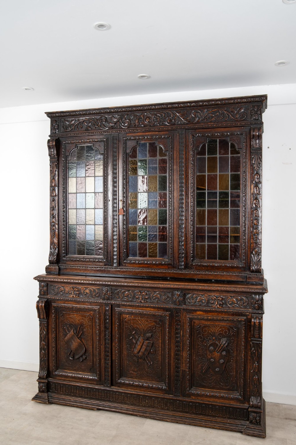 A large late Victorian dark carved oak bookcase featuring three upper glazed doors with coloured leaded glass panels above a lower cupboard base with relief carved emblems. View III