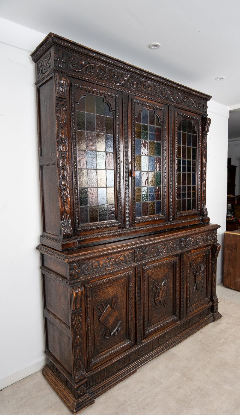 A large late Victorian dark carved oak bookcase featuring three upper glazed doors with coloured leaded glass panels above a lower cupboard base with relief carved emblems. View IV