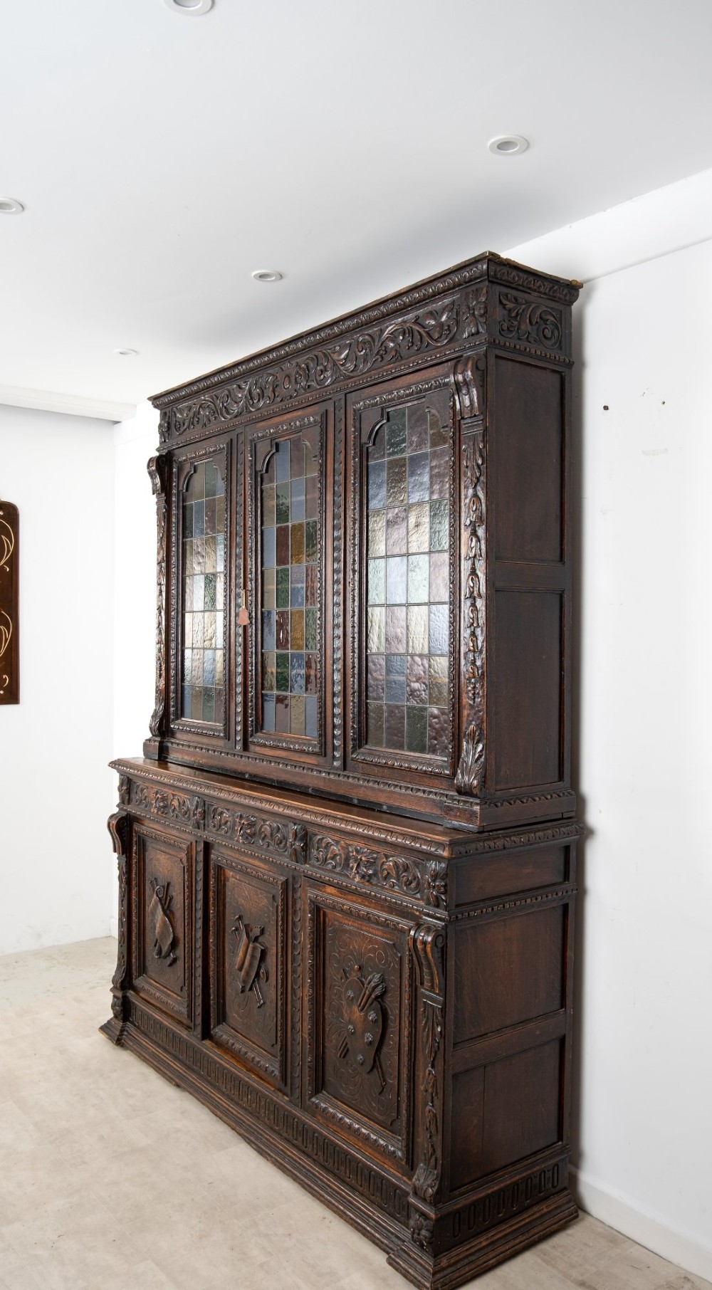 A large late Victorian dark carved oak bookcase featuring three upper glazed doors with coloured leaded glass panels above a lower cupboard base with relief carved emblems. View V