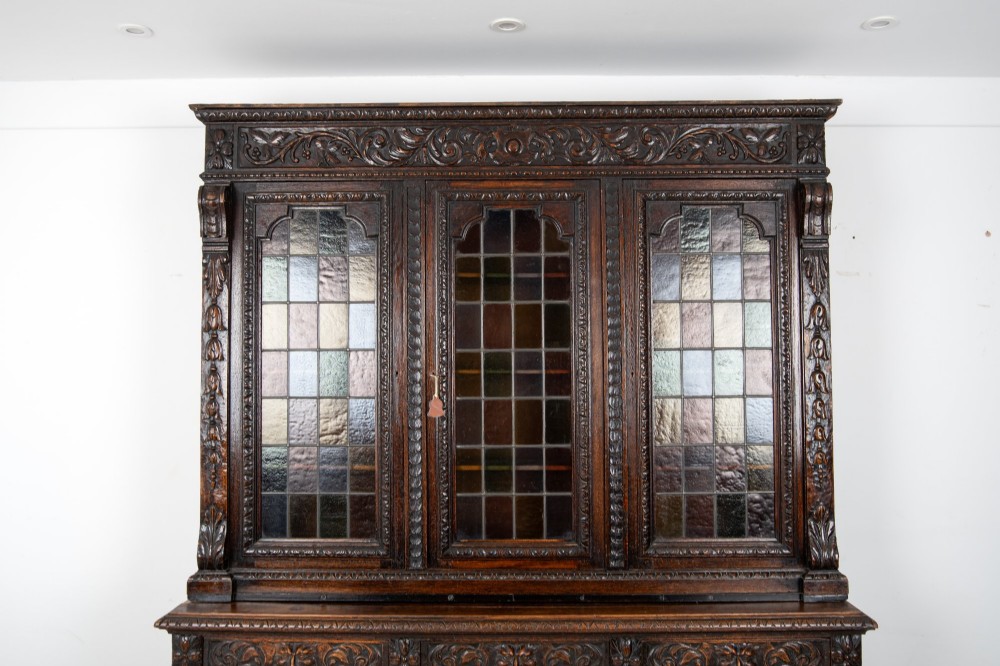 A large late Victorian dark carved oak bookcase featuring three upper glazed doors with coloured leaded glass panels above a lower cupboard base with relief carved emblems. View VI