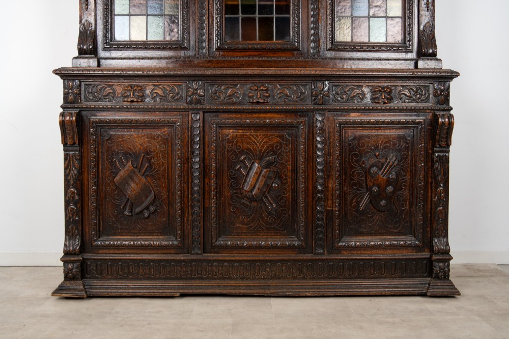 A large late Victorian dark carved oak bookcase featuring three upper glazed doors with coloured leaded glass panels above a lower cupboard base with relief carved emblems. View VII