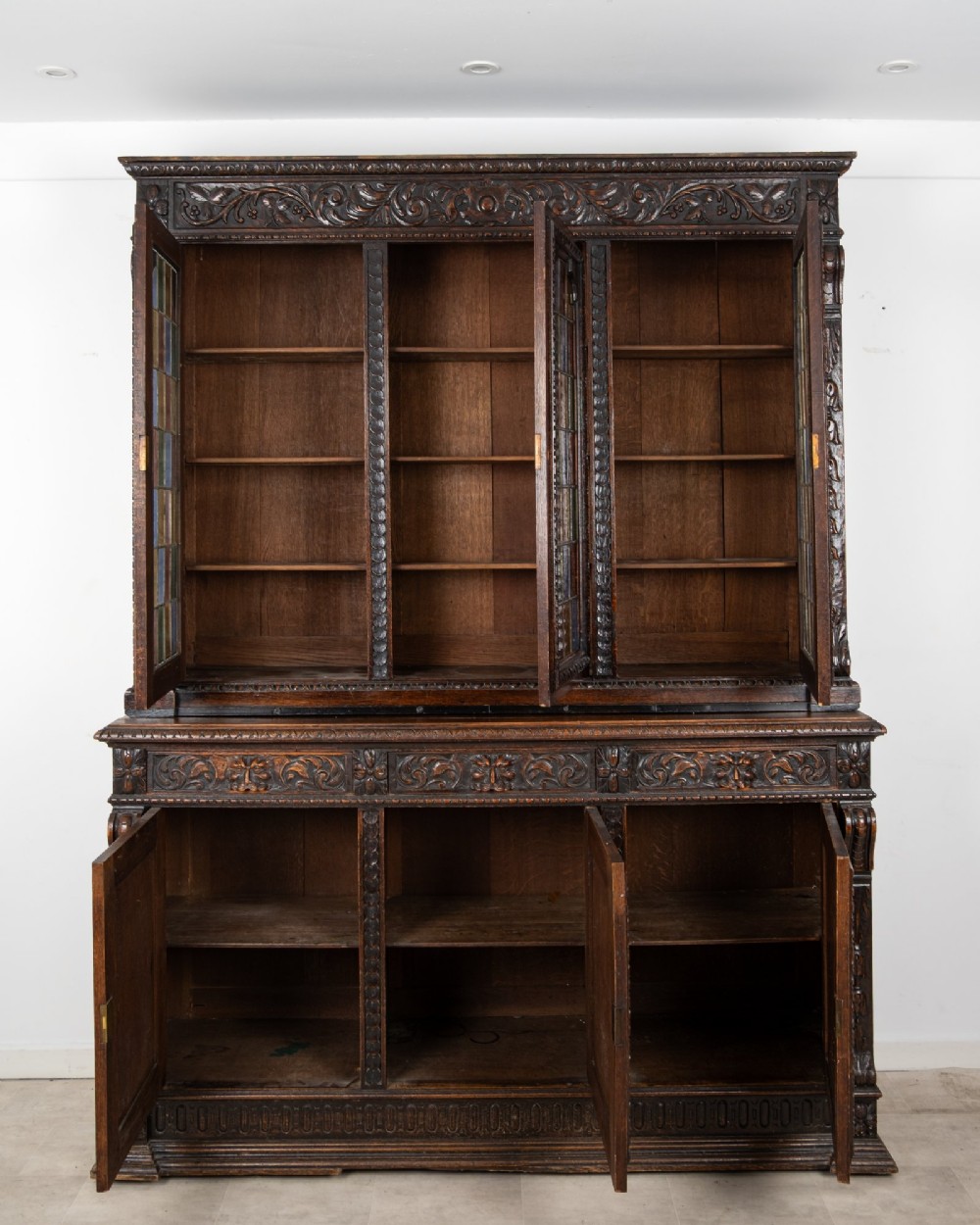 A large late Victorian dark carved oak bookcase featuring three upper glazed doors with coloured leaded glass panels above a lower cupboard base with relief carved emblems. View VIII