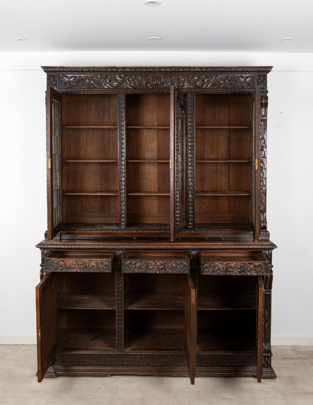 A large late Victorian dark carved oak bookcase featuring three upper glazed doors with coloured leaded glass panels above a lower cupboard base with relief carved emblems. View IX