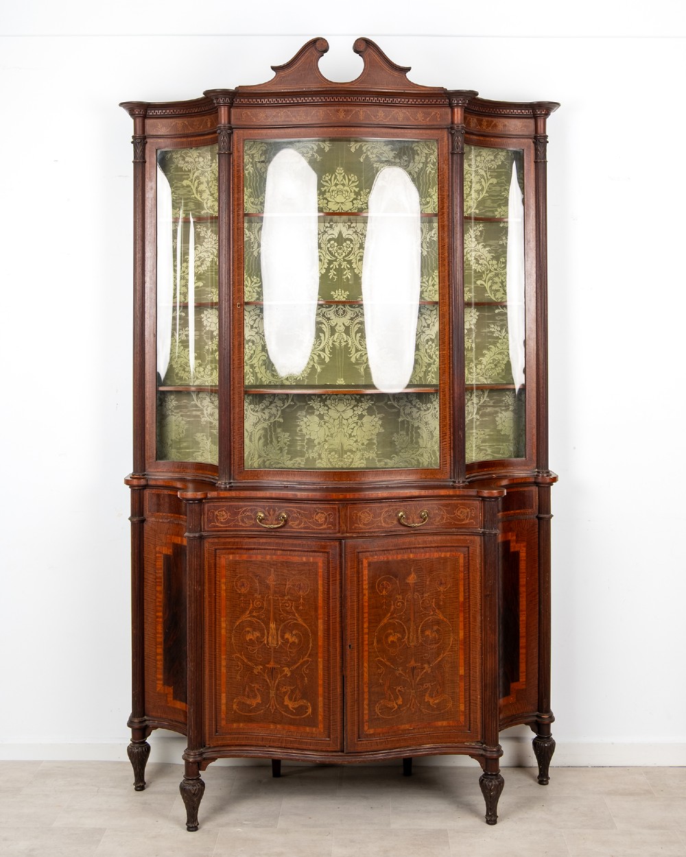 An Edwardian mahogany display cabinet featuring satinwood marquetry inlay, glazed upper doors with green fabric lining, and turned legs.