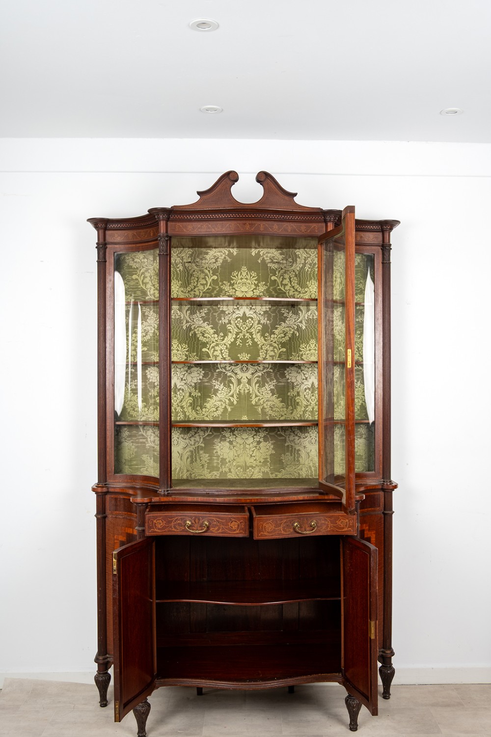 An Edwardian mahogany display cabinet featuring satinwood marquetry inlay, glazed upper doors with green fabric lining, and turned legs. View IX
