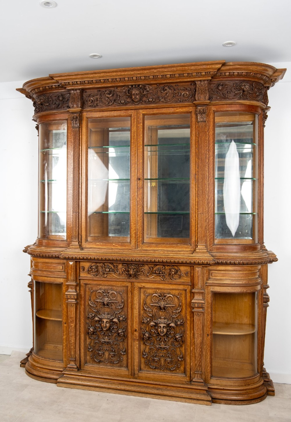 A large Victorian carved oak display cabinet featuring curved glass sides, intricately carved mask motifs on lower door panels, and glass shelving. View II