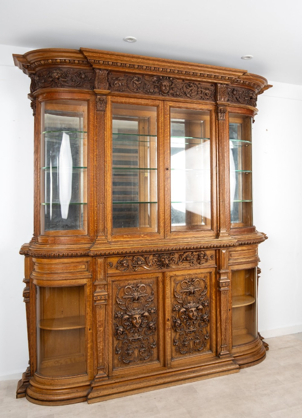 A large Victorian carved oak display cabinet featuring curved glass sides, intricately carved mask motifs on lower door panels, and glass shelving. View III