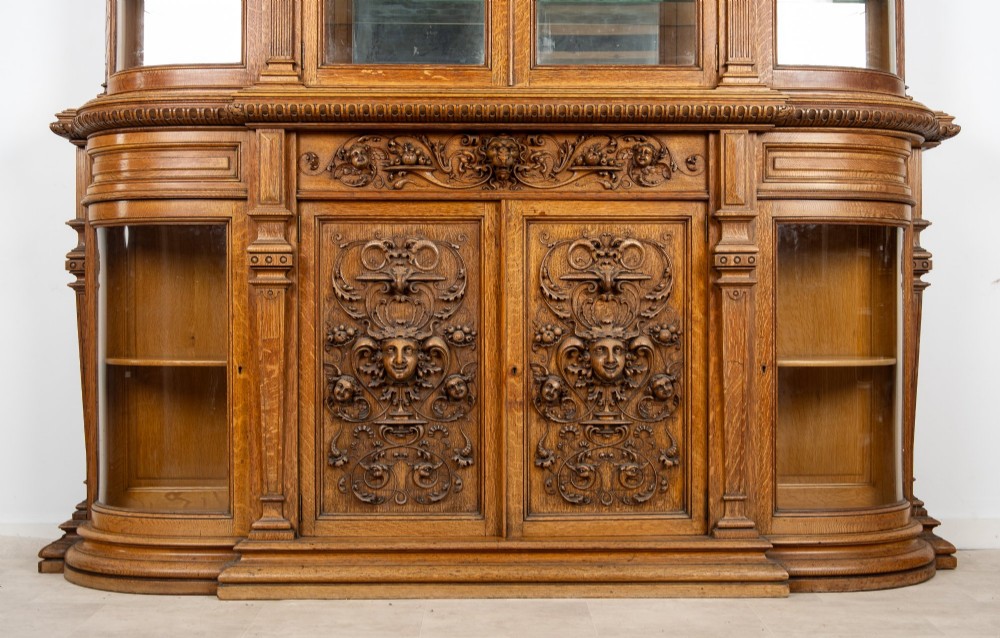 A large Victorian carved oak display cabinet featuring curved glass sides, intricately carved mask motifs on lower door panels, and glass shelving. View IV