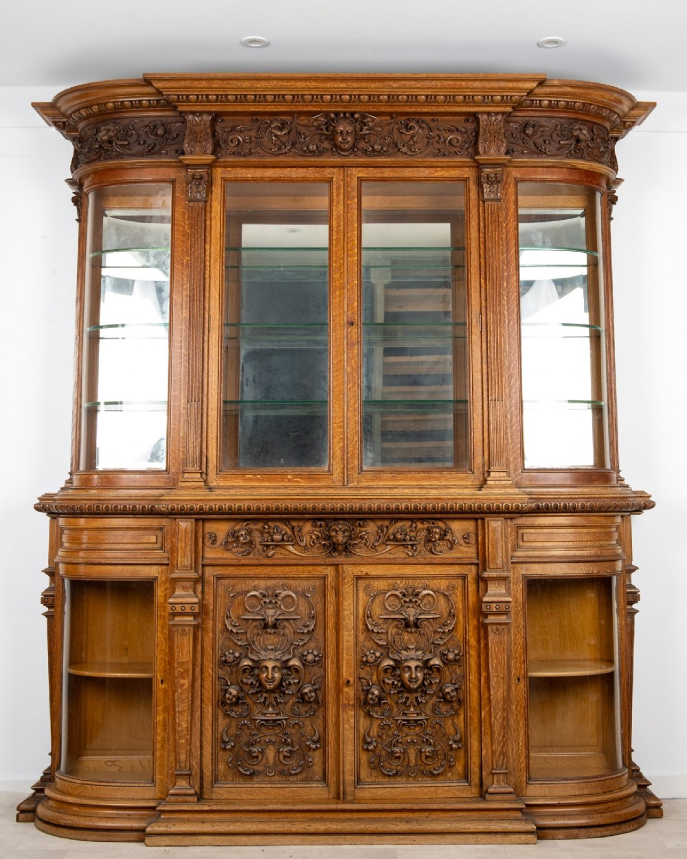 A large Victorian carved oak display cabinet featuring curved glass sides, intricately carved mask motifs on lower door panels, and glass shelving. View V