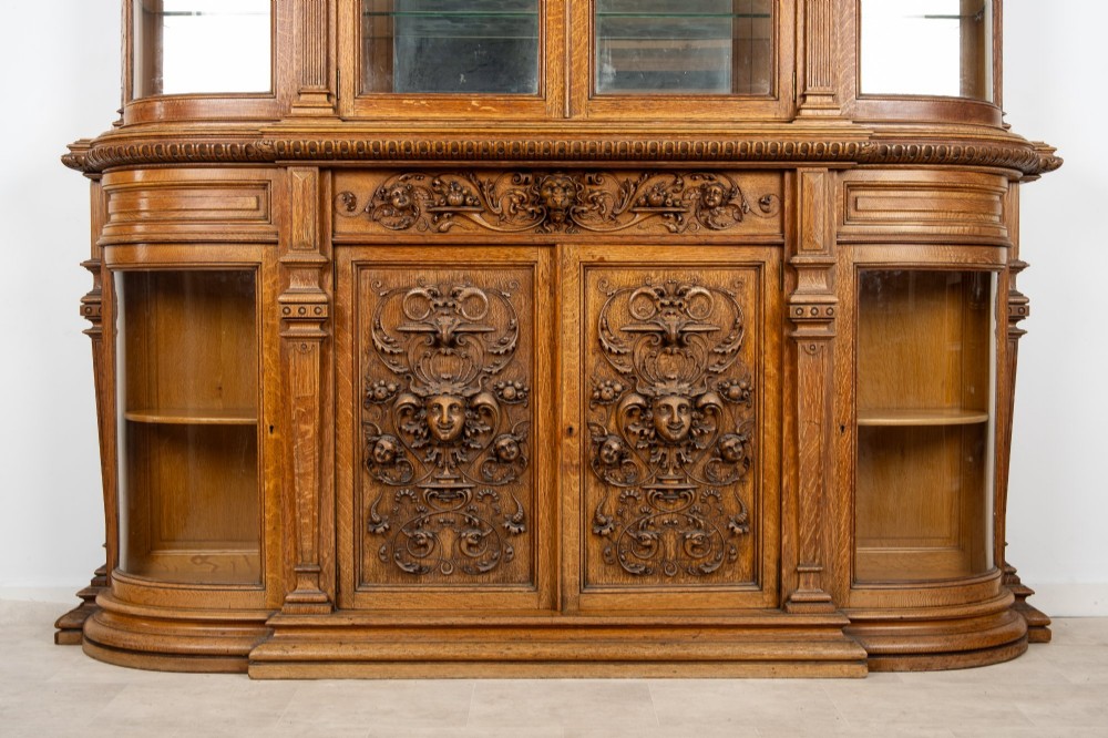 A large Victorian carved oak display cabinet featuring curved glass sides, intricately carved mask motifs on lower door panels, and glass shelving. View VI
