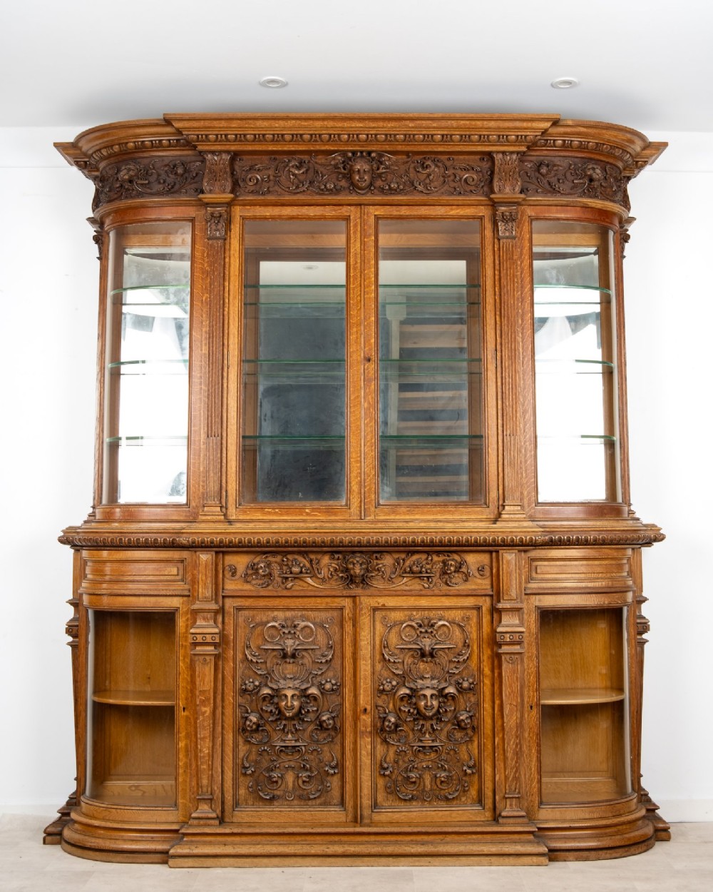 A large Victorian carved oak display cabinet featuring curved glass sides, intricately carved mask motifs on lower door panels, and glass shelving. View VII