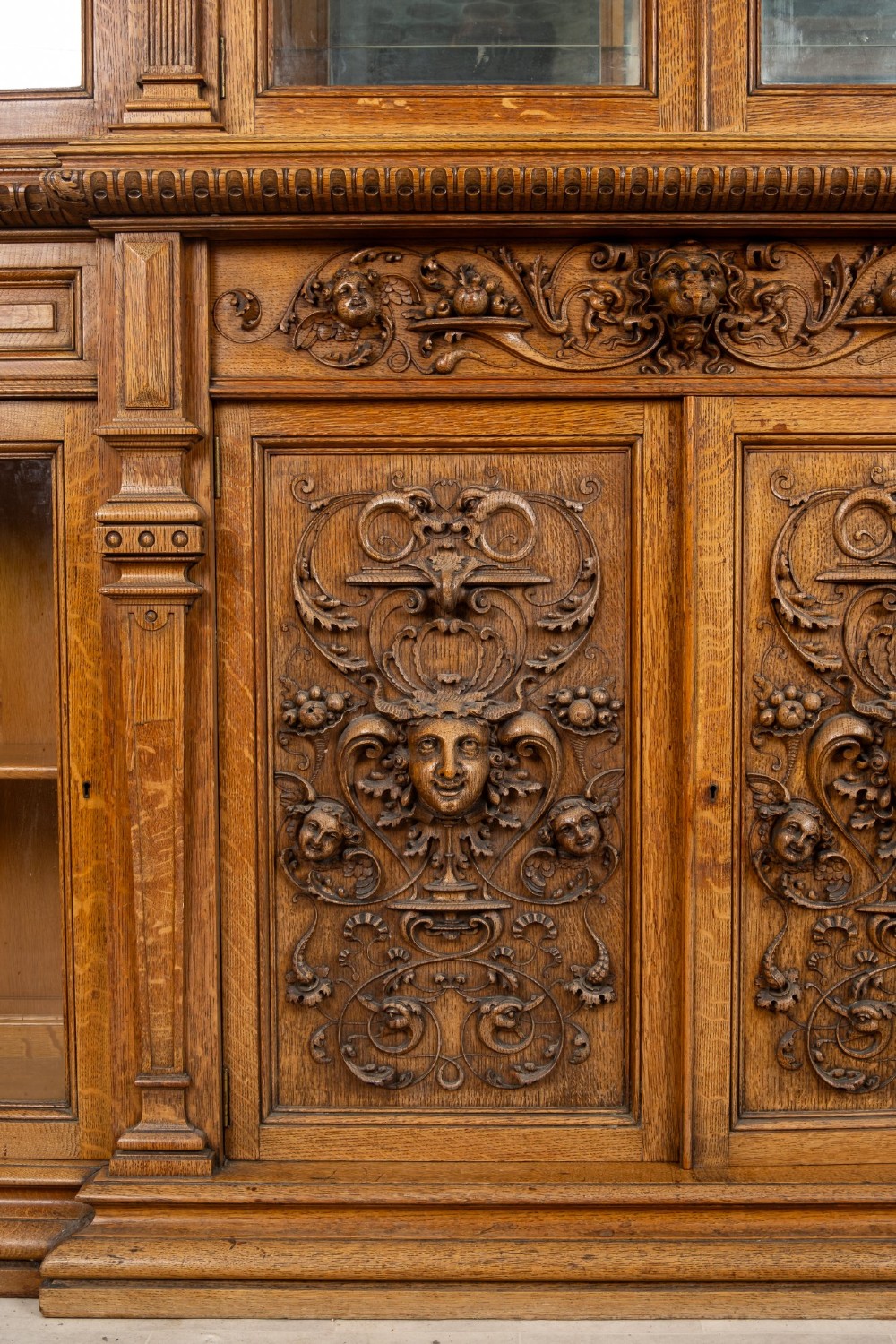 A large Victorian carved oak display cabinet featuring curved glass sides, intricately carved mask motifs on lower door panels, and glass shelving. View VIII