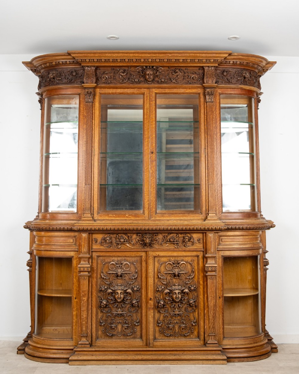 A large Victorian carved oak display cabinet featuring curved glass sides, intricately carved mask motifs on lower door panels, and glass shelving. View IX