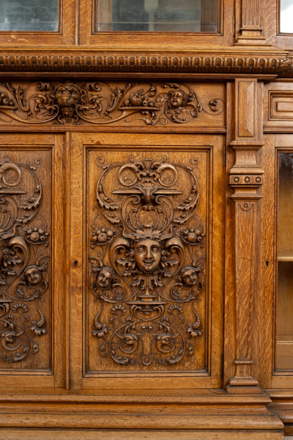 A large Victorian carved oak display cabinet featuring curved glass sides, intricately carved mask motifs on lower door panels, and glass shelving. View X