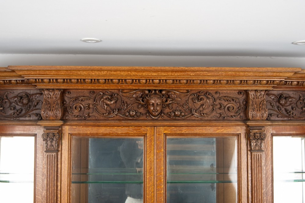 A large Victorian carved oak display cabinet featuring curved glass sides, intricately carved mask motifs on lower door panels, and glass shelving. View XII