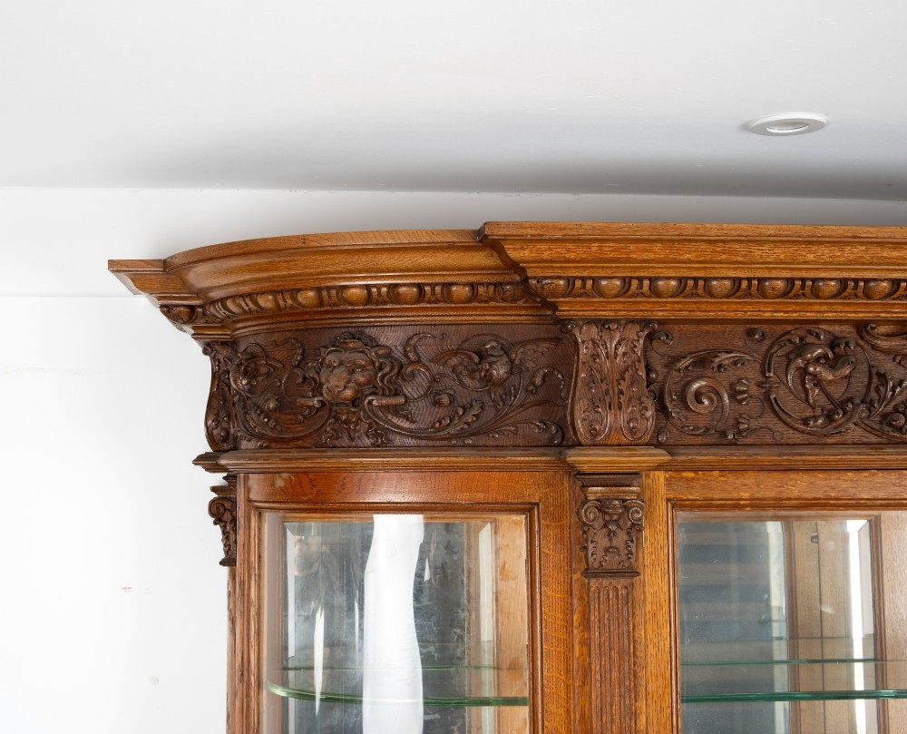 A large Victorian carved oak display cabinet featuring curved glass sides, intricately carved mask motifs on lower door panels, and glass shelving. View XIII