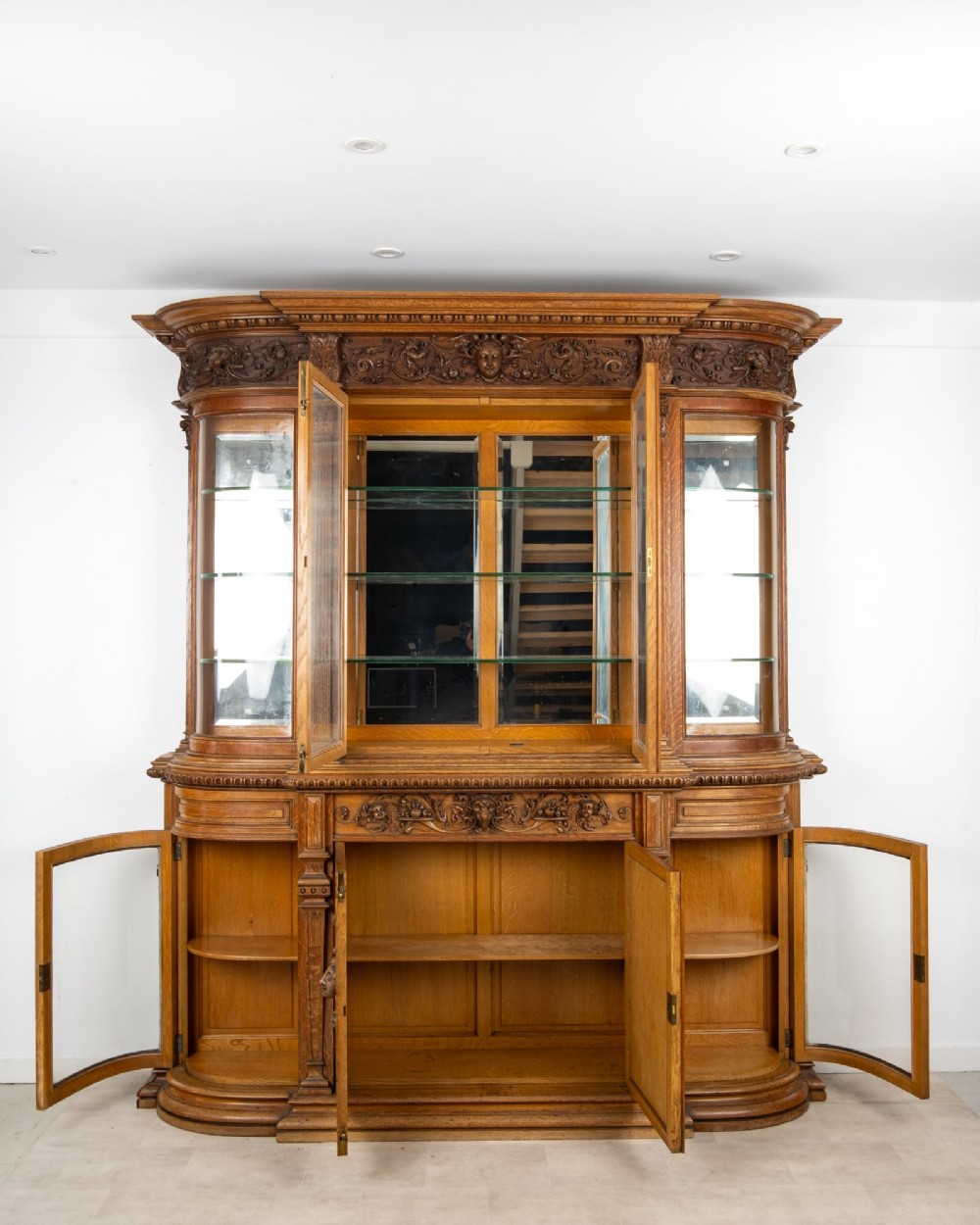 A large Victorian carved oak display cabinet featuring curved glass sides, intricately carved mask motifs on lower door panels, and glass shelving. View XV