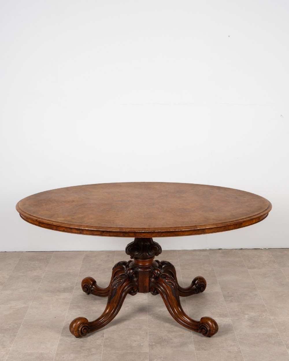 A Victorian oval burr walnut loo table tilted vertically to display symmetrical bookmatched grain patterns above a carved pedestal base with four scrolled legs. View II