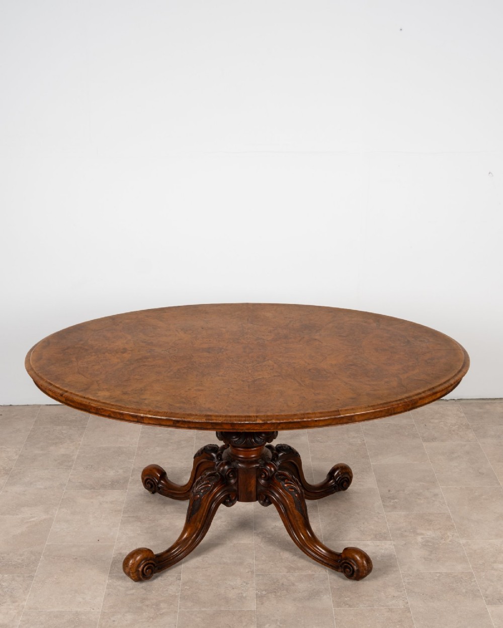 A Victorian oval burr walnut loo table tilted vertically to display symmetrical bookmatched grain patterns above a carved pedestal base with four scrolled legs. View III