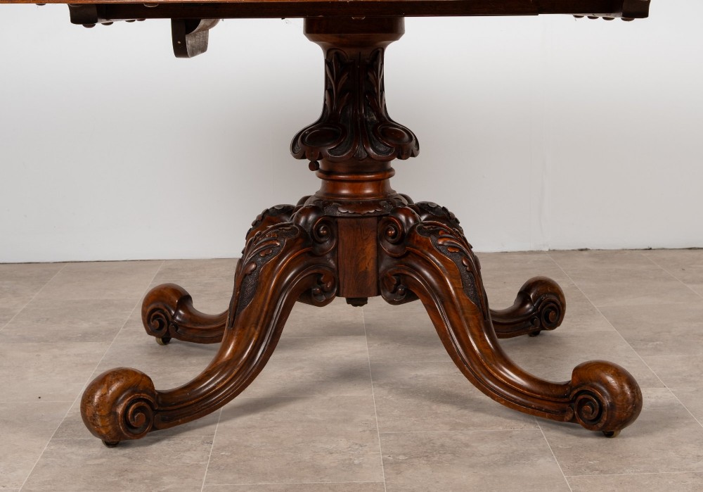 A Victorian oval burr walnut loo table tilted vertically to display symmetrical bookmatched grain patterns above a carved pedestal base with four scrolled legs. View IV