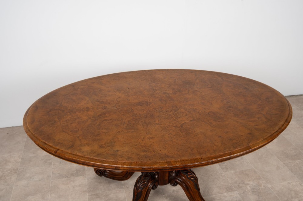 A Victorian oval burr walnut loo table tilted vertically to display symmetrical bookmatched grain patterns above a carved pedestal base with four scrolled legs. View V