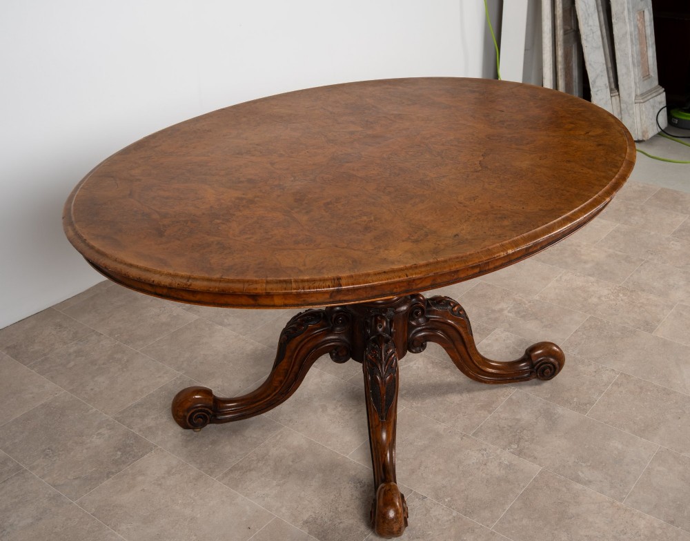 A Victorian oval burr walnut loo table tilted vertically to display symmetrical bookmatched grain patterns above a carved pedestal base with four scrolled legs. View VI