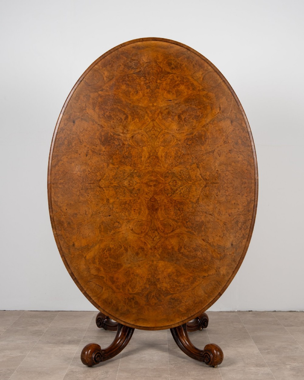 A Victorian oval burr walnut loo table tilted vertically to display symmetrical bookmatched grain patterns above a carved pedestal base with four scrolled legs.
