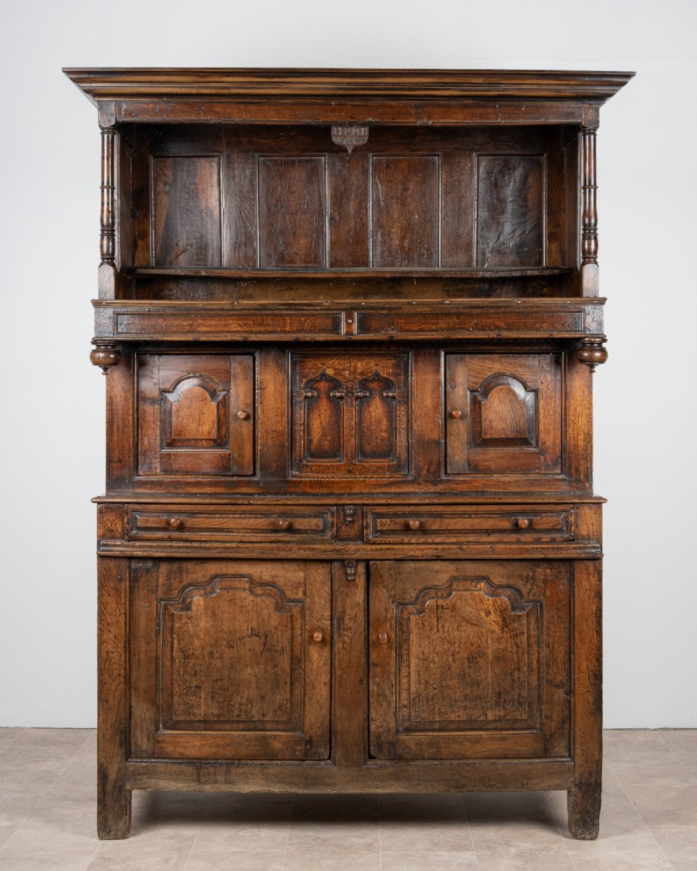A dark oak three-tier Welsh cwpwrdd tridarn featuring an open upper canopy, middle cupboard section with raised arch panels and turned pillars, and a base cupboard with two drawers.