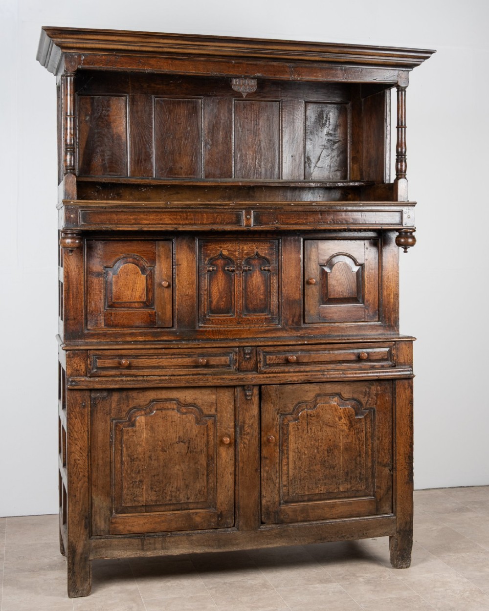 A dark oak three-tier Welsh cwpwrdd tridarn featuring an open upper canopy, middle cupboard section with raised arch panels and turned pillars, and a base cupboard with two drawers. View II