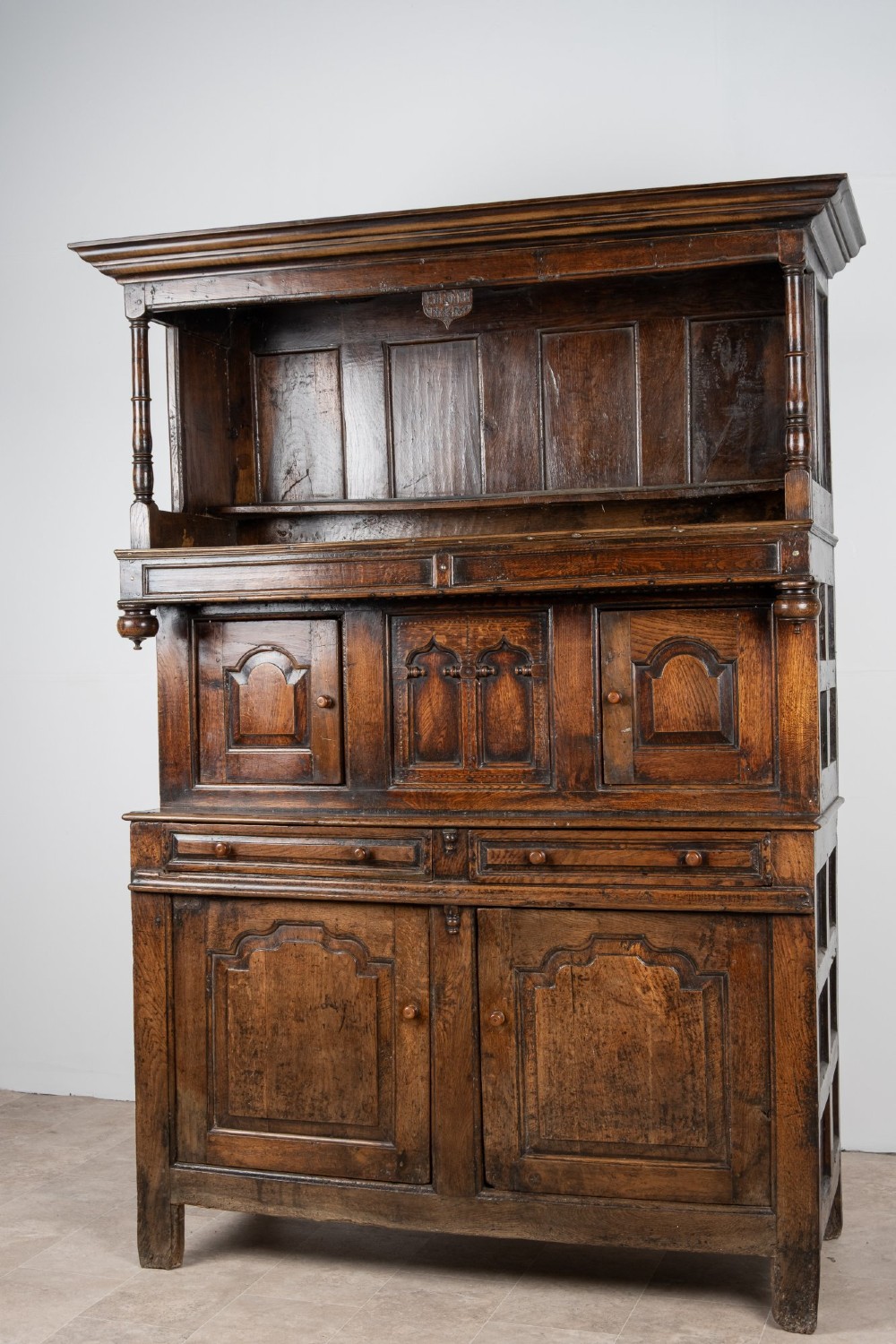 A dark oak three-tier Welsh cwpwrdd tridarn featuring an open upper canopy, middle cupboard section with raised arch panels and turned pillars, and a base cupboard with two drawers. View V
