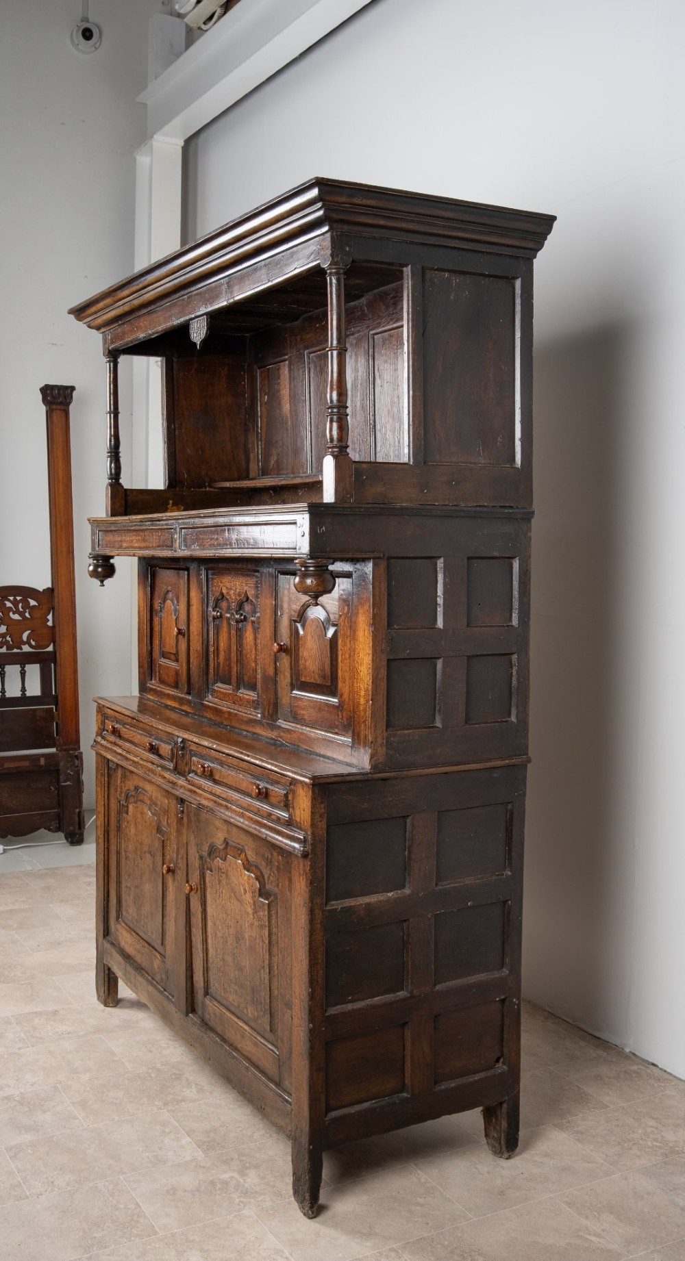 A dark oak three-tier Welsh cwpwrdd tridarn featuring an open upper canopy, middle cupboard section with raised arch panels and turned pillars, and a base cupboard with two drawers. View VI
