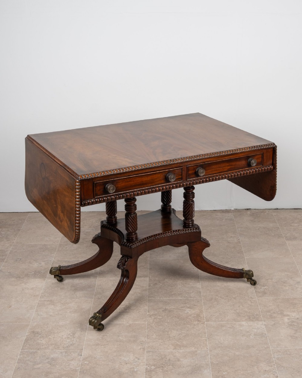 An antique Regency mahogany drop-leaf sofa table with two drawers, spiral-turned column supports, and splayed legs on brass castors.