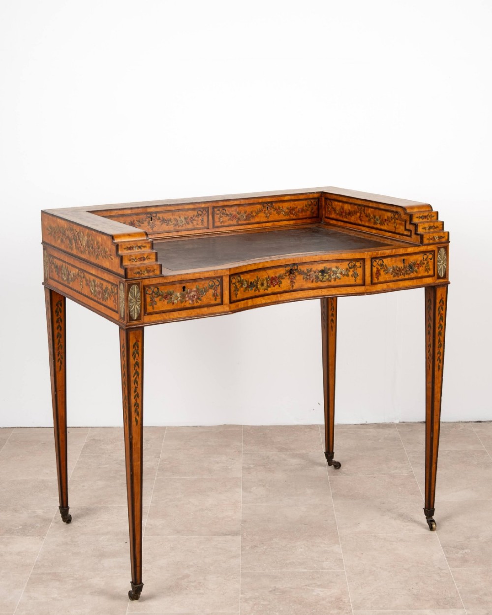 A warm gold satinwood Carlton House writing desk featuring hand-painted floral swags, a dark leather writing surface, and tapered legs on brass castors.